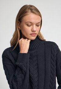 Mörkgrå stickad tröja med hög krage, kabelstickad mönster och ribbstickade ärmslut, med en strukturerad och kraftig design.