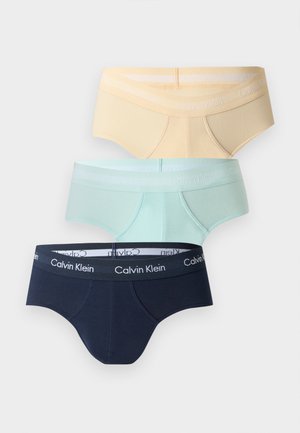 Athletischer dunkelhäutiger Mann, barfuß stehend, trägt marineblaue Calvin Klein Unterwäsche vor einfachem weißem Hintergrund.