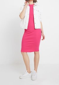 Upnuté růžové žebrované šaty s kulatým výstřihem, v kombinaci s bílou džínsovou bundou a bílými teniskami, postavené proti jednobarevnému pozadí.