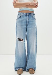 Hellblaue weit geschnittene Jeans mit einem aufgerissenen Knie. Der Stoff sieht aus wie Denim, hat eine ausgewaschene Optik und einen lockeren Schnitt.