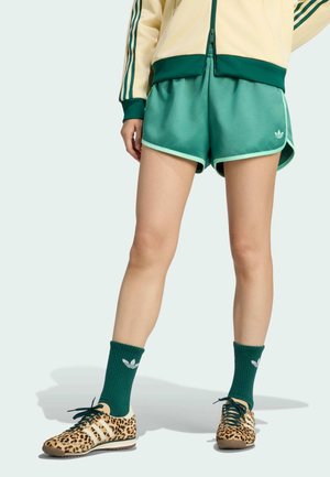 Person iført grønne atletikshorts, mørkegrønne sokker med et logo, leopardmønstrede sneakers og en lyse jakke med grønne striber.
