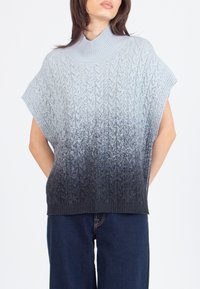 Žena u bezrukavnoj plavoj ombre pletenoj jumperici s lažnim ovratnikom i tamnoplave hlače ispred jednostavne pozadine.