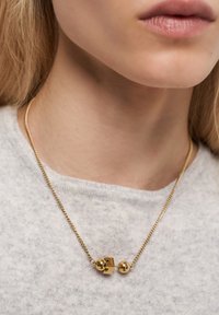 Nærbillede af en guldkæde med et unikt sfærisk og rektangulært lås, båret af en person i en lysegrå top.