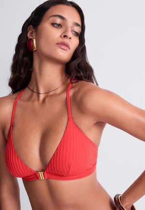 Femme aux longs cheveux ondulés portant un haut de bikini côtelé rouge, de grandes boucles d'oreilles rouges et dorées, et un fin collier en or sur un fond uni.