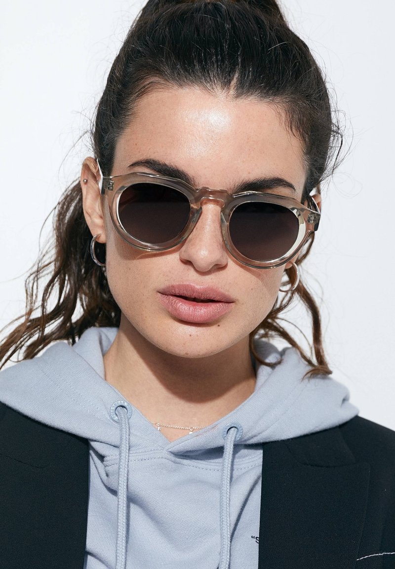 Ümarad prillid, millel on poolläbipaistev helepruun raam ja tumedad läätst. Kujunduses on paksud servad ja metallist aktsendid hinge juures.