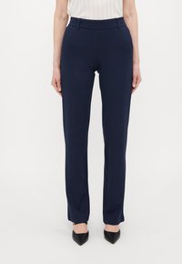 Navy blue, tailored trousers with a smooth fabric, straight fit, belt loops, and minimal stitching detail. Paired with black, pointed-toe heels.