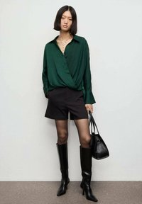 Blusa de seda verde con diseño envolvente, pantalones cortos negros a medida, botas altas negras, collar de cadena de plata y un pequeño bolso negro.