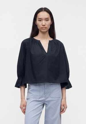 Femme aux longs cheveux noirs portant un chemisier noir ample à col en V et un pantalon taille haute bleu clair, debout devant un fond uni.
