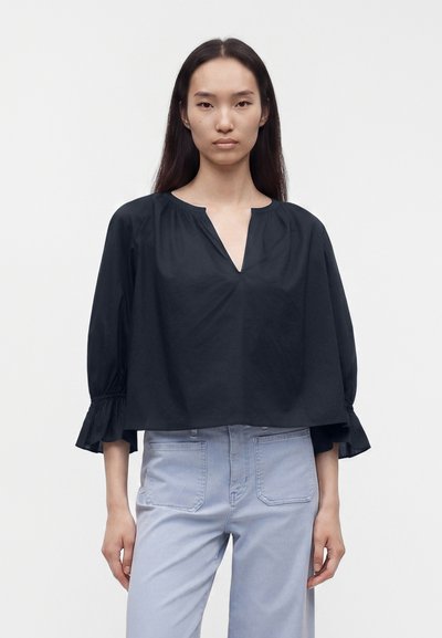 Femme aux longs cheveux noirs portant un chemisier noir ample à col en V et un pantalon taille haute bleu clair, debout devant un fond uni.