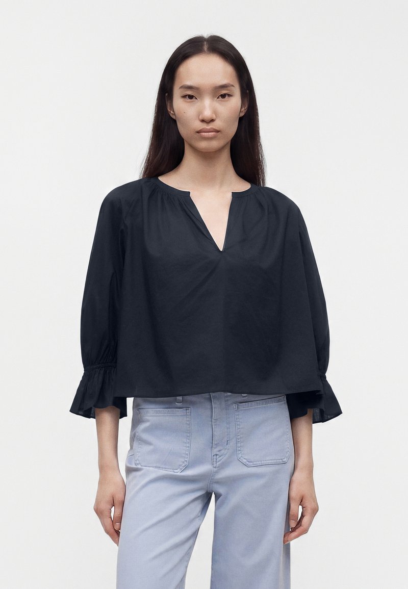 Femme aux longs cheveux noirs portant un chemisier noir ample à col en V et un pantalon taille haute bleu clair, debout devant un fond uni.