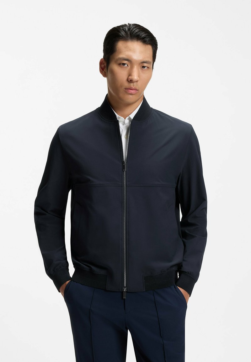 Homme aux cheveux foncés portant une veste noire ajustée à fermeture éclair sur une chemise blanche, les mains dans les poches d’un pantalon bleu marine, sur un fond uni.
