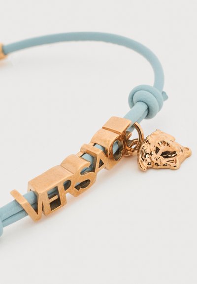 Βραχιόλι από ανοιχτό μπλε κορδόνι με γράμματα σε χρυσή απόχρωση που σχηματίζουν τη λέξη "VERSACE" και μια μικρή χρυσή μινιατούρα κεφαλής Μέδουσας.