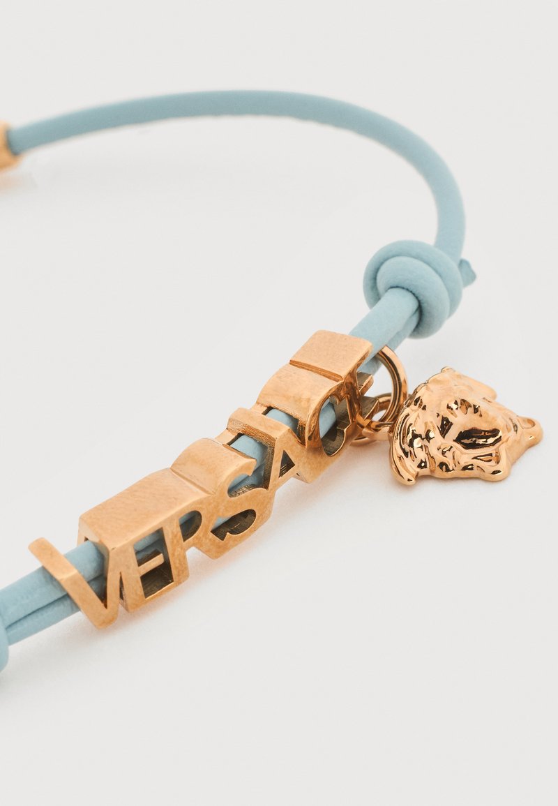 Light blue cord bracelet with gold-tone letters spelling "VERSACE" and a small gold-tone Medusa head charm.