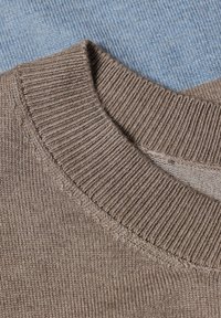 Gros plan sur le col d'un pull en tricot beige avec une texture côtelée posé sur un fond en tissu bleu clair.