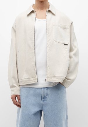 Person iført en lysebeige lynlåstrøje med en brystlomme, hvid t-shirt og lyseblå cowboybukser mod en hvid baggrund.