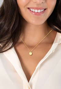 Arany nyaklánc kör alakú medállal, vékony csavart lánccal, fehér blúzra fektetve, sima bőrfelület háttér előtt.