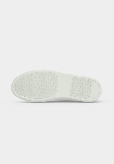 Semelle en caoutchouc blanc d’une chaussure avec des motifs de grip texturés, vue par dessous sur un fond gris clair uni.