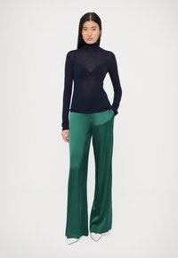 Granatowy, długi rękaw golf z dopasowanym krojem, zestawiony z zielonymi, szerokimi spodniami z gładkiej tkaniny. Model stoi z rękami w kieszeniach.