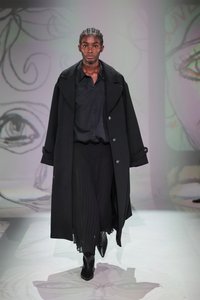 Čierny trenčkot prehodený cez čiernu košeľu s gombíkmi a plisované nohavice; model má na sebe priliehavé čierne členkové topánky; minimalistický, monochromatický dizajn.