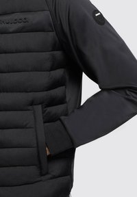 Gilet imbottito nero con una parte superiore in tessuto liscio e una parte inferiore imbottita testurizzata. Presenta una tasca laterale e un logo sul petto e sulla manica.