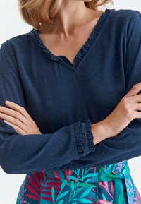 Blusa navy con scollatura a V e colletto a volant. Le maniche lunghe hanno dettagli a volant. La modella indossa una gonna colorata e a fantasia.