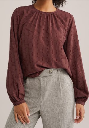 Kobieta w bordowej, teksturowanej bluzce z długim rękawem, włożonej do szarych spodni w jodełkę z wysokim stanem i zapięciem na guziki.