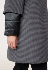 Šedý vzorovaný kabát s černým prošívaným rukávem. Hladký materiál a přiléhavý střih, s mírně rozšířeným lemem a rukou s prsteny.