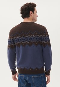 Brun och blå tröja med geometriska mönster, stickad textur, rund halsringning och ribbad mudd och fåll. Har en kontrasterande design på oket.