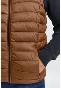 Brauner, gepolsterter Weste mit gestepptem Design und schwarzem Reißverschluss, getragen über einem navy-blauen Pullover. Sie hat seitliche Taschen und eine glatte Textur.