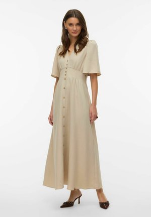 Donna che indossa un abito lungo beige con bottoni frontali, maniche corte a sbuffo e tacchi marroni a punta, in piedi su sfondo bianco.