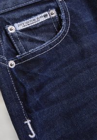 Σκούρα τζιν από τζιν με ξεφτισμένες λεπτομέρειες και λευκό ράψιμο. Η ετικέτα φέρει την επιγραφή «JUICY COUTURE JEANS». Ασημένια μεταλλικά στοιχεία στις τσέπες.