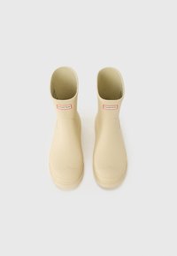 Bottes en caoutchouc beige à bout arrondi et tige courte, avec un label logo rouge et blanc à l'avant. Texture lisse, sans motifs.