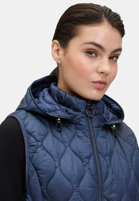 Námořnický prošívaný vest s vysokým límcem a kapucí. Obsahuje centrální zip a detaily s tkaničkami. Vyrobeno z hladké, texturované tkaniny.