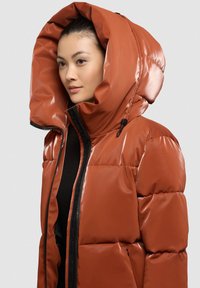 Blizganti, degintos oranžinės spalvos pūkinė striukė su dideliu gobtuvu, priekyje užtrauktuku ir elastingomis raištelėmis; turi dygsniuotą raštą ir juodus akcentus.