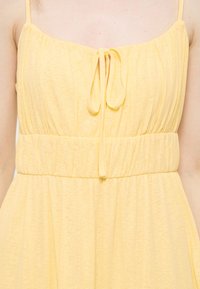 Robe jaune sans manches avec fines bretelles, poitrine froncée, taille élastique et petit nœud noué au centre devant.