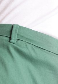 Tissu vert des pantalons, présentant une texture lisse et une taille cousue. En arrière-plan, une chemise blanche est visible en dessous.