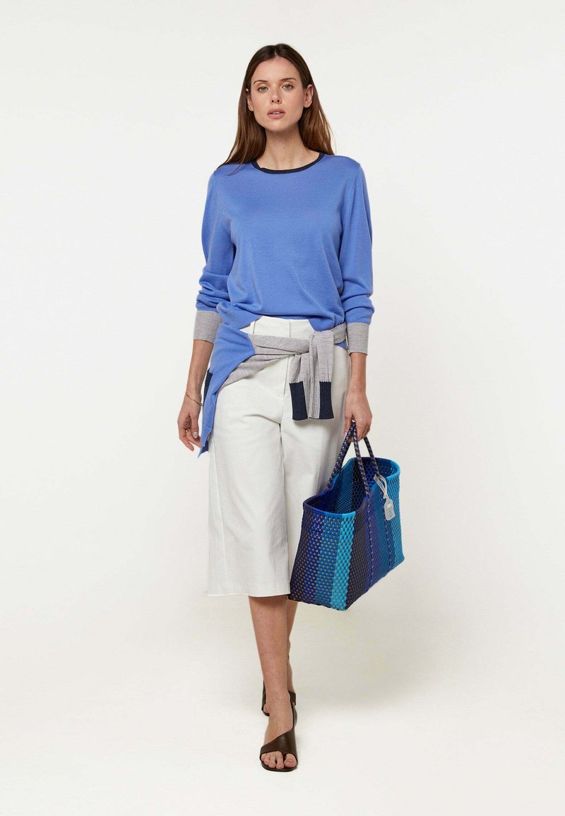 Frau trägt blaues Langarmshirt, weißen knielangen Rock, grauen Pullover um die Taille gebunden, hält blaue Tragetasche und geht vorwärts vor weißem Hintergrund.