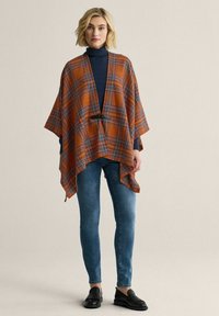 Orange og blå ternet poncho med åben front, bælte i taljen og en løs, flydende design. Sammen med en navy turtleneck og jeans.