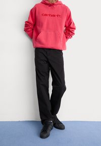 Felpa rossa in cotone con tasca a marsupio frontale e logo, abbinata a pantaloni neri e sneakers nere con superfici testurizzate.