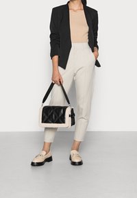 Černá prošívaná kabelka s krémovým umělým kožíškem, kterou drží osoba v béžovém topu a světlých kalhotách, obutá do krémových mokasín s tmavými podrážkami.