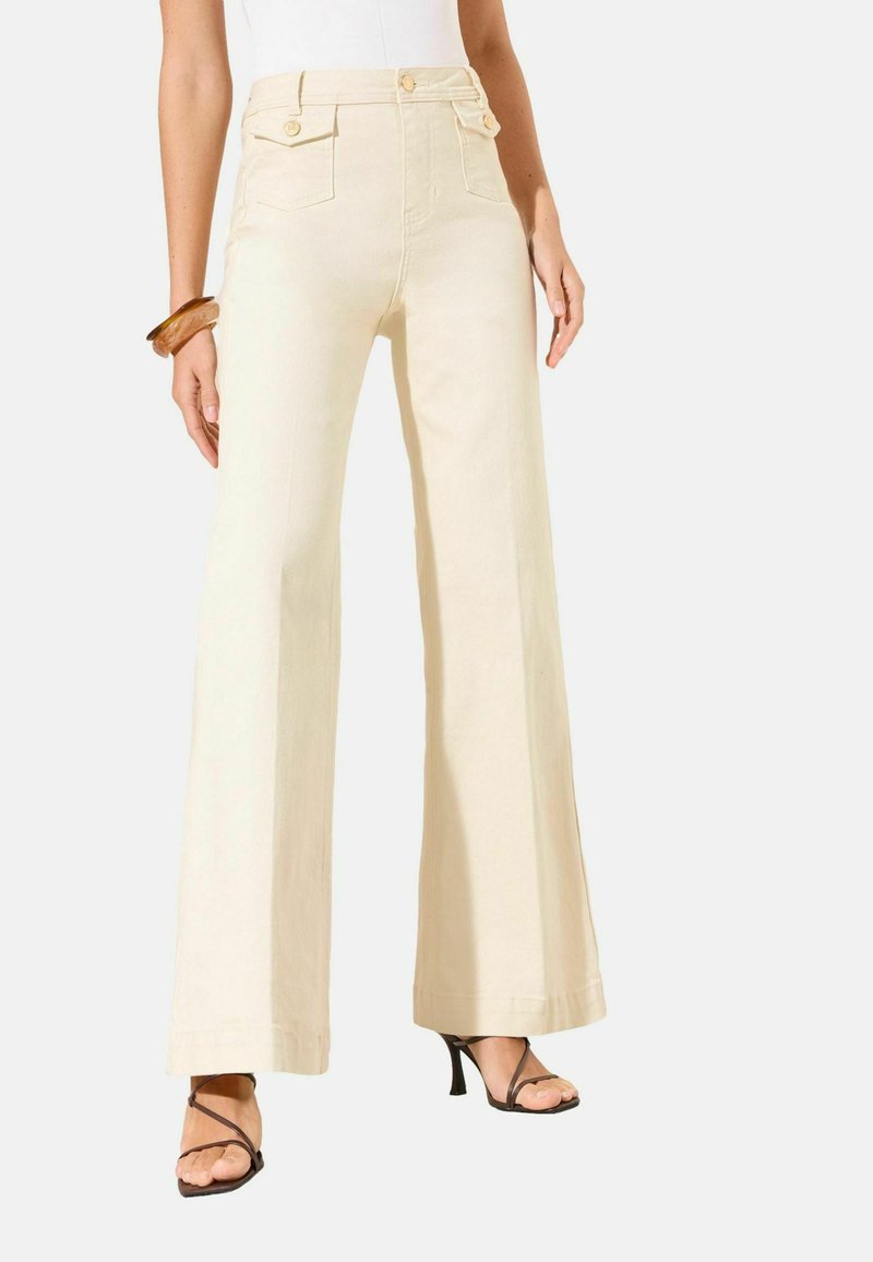 Žena nosi krémové nohavice s vysokým pásom a širokými nohavicami, s predným zapínaním na gombíky a vreckami, v kombinácii s hnedými sandálmi na podpätku a dreveným náramkom.