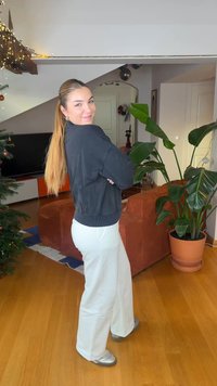 Melna bumbvedējspaka ar ribveida apakšmalu, kopā valkātas baltas platu kāju bikses. Pelēkas kurpes un gari, taisni mati. Redzami zaļi augi un brūnas mēbeles.