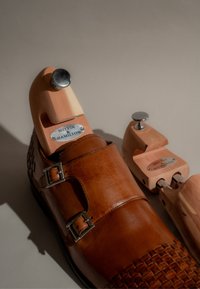 Braune Lederschuhe mit zwei Schnallen und geflochtenem Design; Holzschuhspanner mit metallischen Akzenten, beschriftet mit "Melvin & Hamilton."