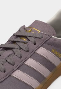 Κοντινό πλάνο μοβ σουέντ παπουτσιού Adidas με ροζ ρίγες, γκρι κορδόνια, χρυσή επιγραφή "München" και καουτσούκ σόλα.