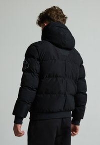 Doudoune noire avec capuche, présentant des sections matelassées et un patch logo sur la manche gauche. Tissu lisse avec une finition mate.