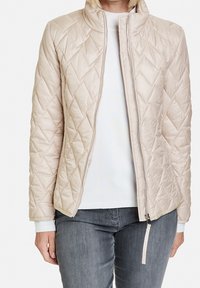 Beige quiltet jakke med høj krave, frontlynlås og hvide manchetter. Det strukturerede stof og diamantmønsteret forbedrer designet.