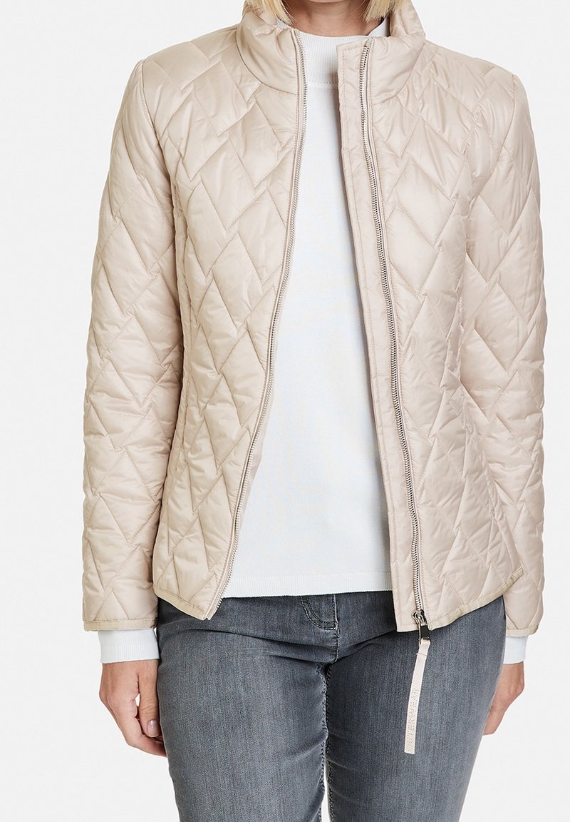 Beige quiltet jakke med høj krave, frontlynlås og hvide manchetter. Det strukturerede stof og diamantmønsteret forbedrer designet.