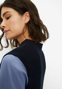 Marineblauer Strickpullover mit strukturiertem Finish und hohem Kragen, über einem hellblauen Hemd getragen. Das Design zeichnet sich durch ärmelloses Styling und einen gerippten Saum aus.