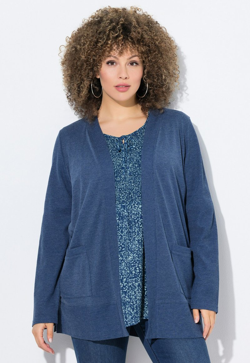 Mörkblå cardigan i texturerad tyg, med öppen framsida, två framfickor, bärs över en blus med blommönster.