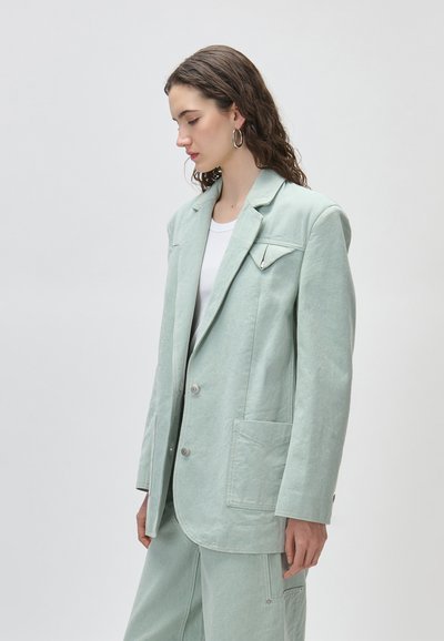 Νεαρή γυναίκα με κυματιστά μαλλιά, που φοράει μεγάλα κρίκους σκουλαρίκια και κοστούμι σε μέντα πράσινο χρώμα, με σακάκι και παντελόνι πάνω από λευκό πουκάμισο, κοιτάζοντας προς τα αριστερά.